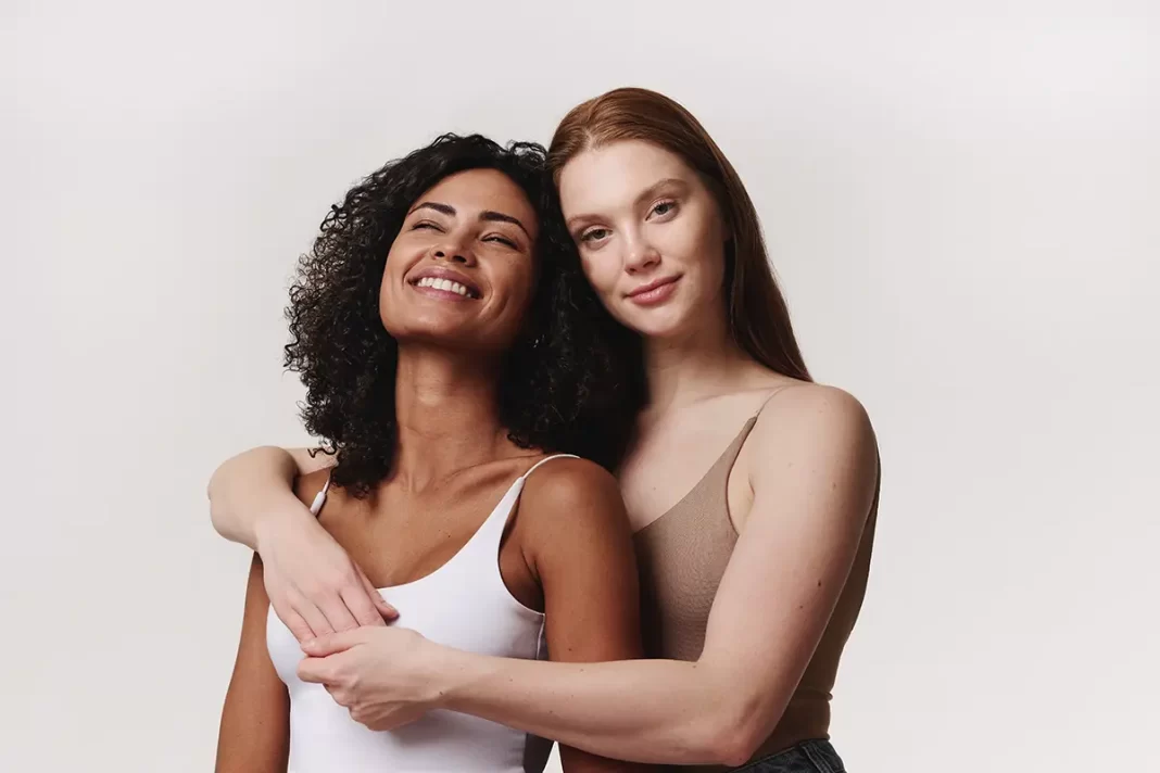 Dos mujeres, una negra y pelo chino y una blanca de pelo castaño claro, ambas con blusas de tirantes y piel radiante abrazándose.
