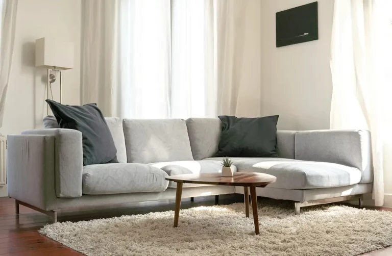 Sala moderna con sofá modular gris claro, cojines oscuros y mesa de centro de madera sobre alfombra blanca