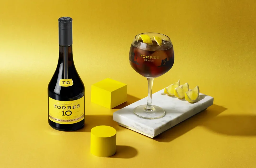 torres-ruta-del-legado-cdmx-cena-gratis Botella de Torres 10 Reserva Imperial junto a cóctel con hielo y limón sobre fondo amarillo de estilo minimalista.